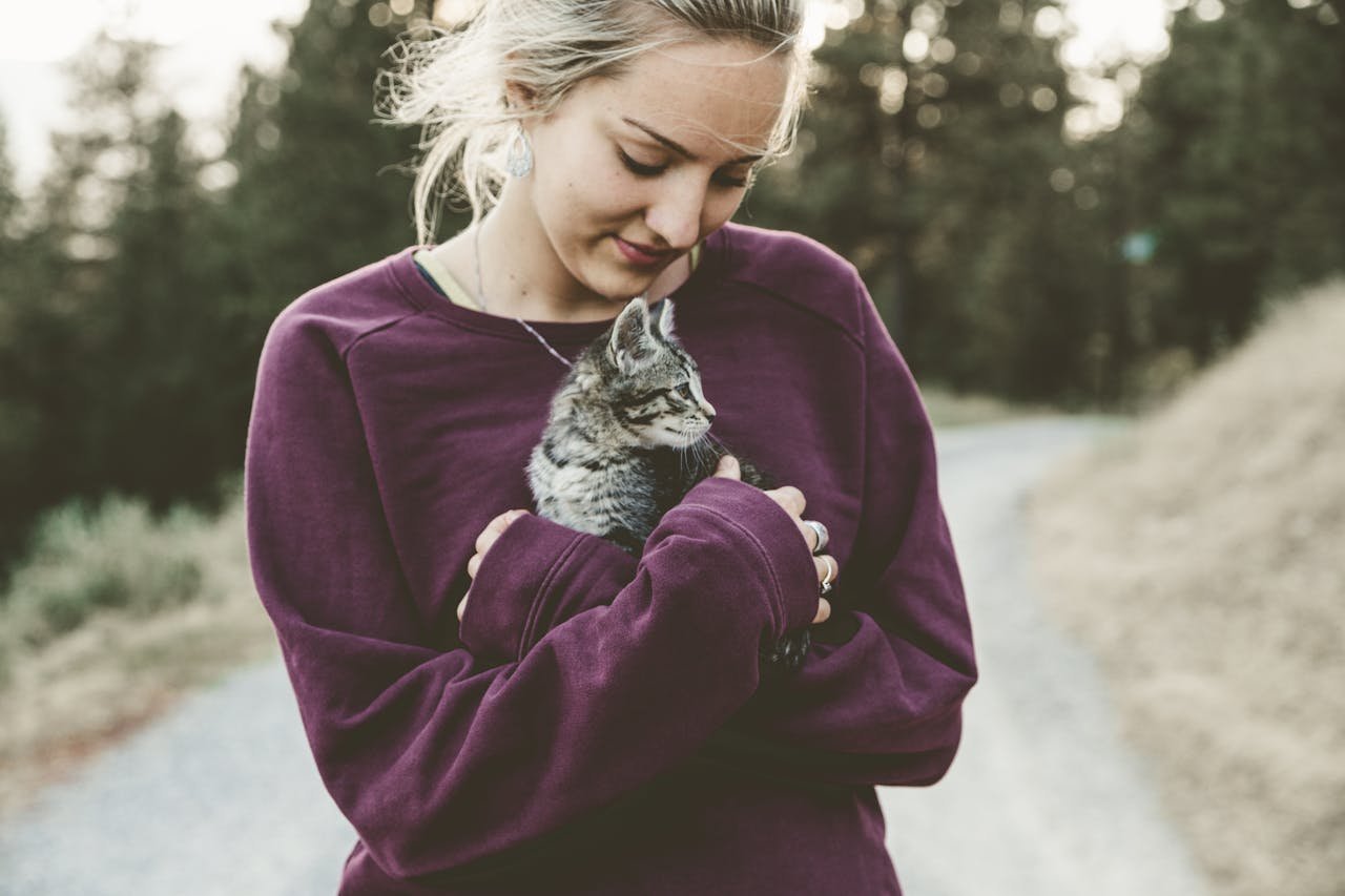 Young woman in a purple sweater smiling down at a kitten shes holding outdoors on a pathway.